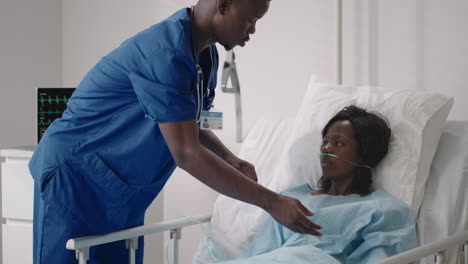 Mujer-Afroamericana-Descansando-En-La-Cama-Del-Hospital-Después-De-La-Cirugía-Hablando-Con-Un-Joven-Enfermero.-Retrato-De-Una-Enfermera-Afroamericana-Que-Ayuda-A-Una-Paciente-Enferma-Acostada-En-La-Cama
